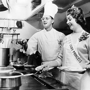 Chef and Dairy Princess in a kitchen, wearing chef hat and crown, smiling while cooking.