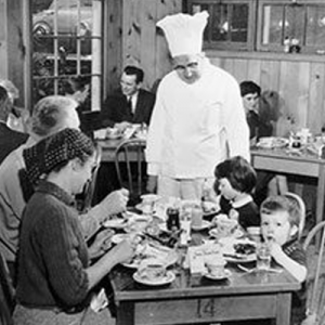 Chef in white uniform serving diners at a crowded restaurant table. Black and white photo.