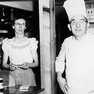Two people, a woman and a chef, stand in a restaurant doorway. Woman holds a notepad, chef smiles, wearing a hat.