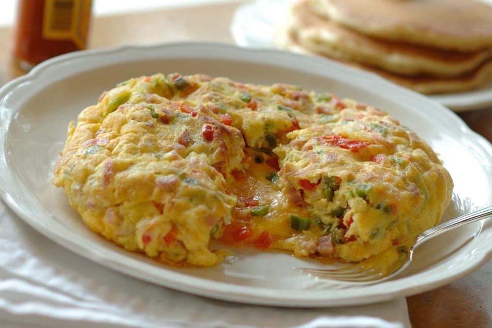 Omelet on a white plate, filled with red peppers, green peppers, and cheese. Stack of pancakes in background.