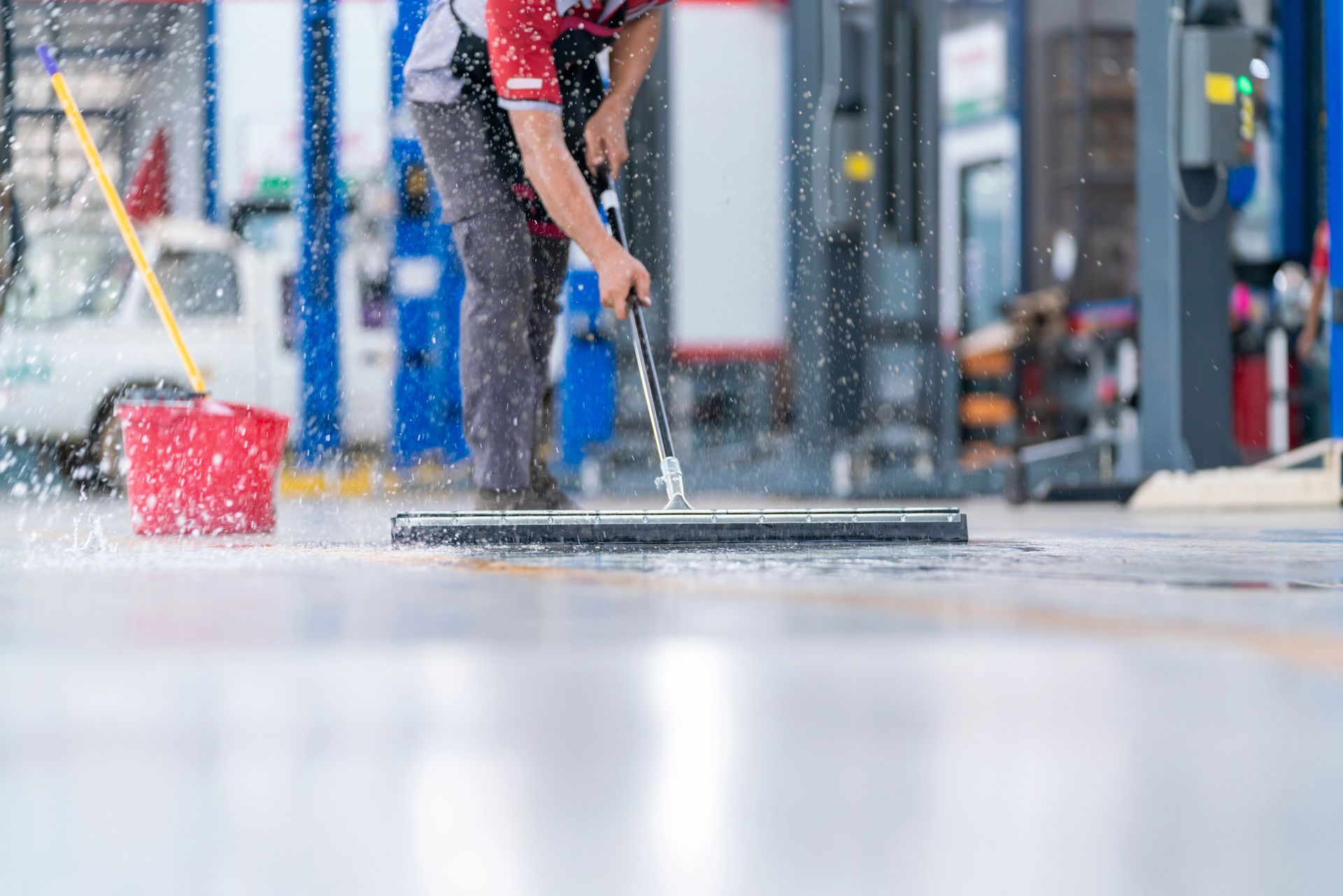 Person mopping a wet garage floor with a squeegee; a red bucket and a vehicle are in the background.