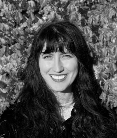 A smiling person with dark, wavy hair, shown from the chest up against a background of foliage in a black-and-white photo.
