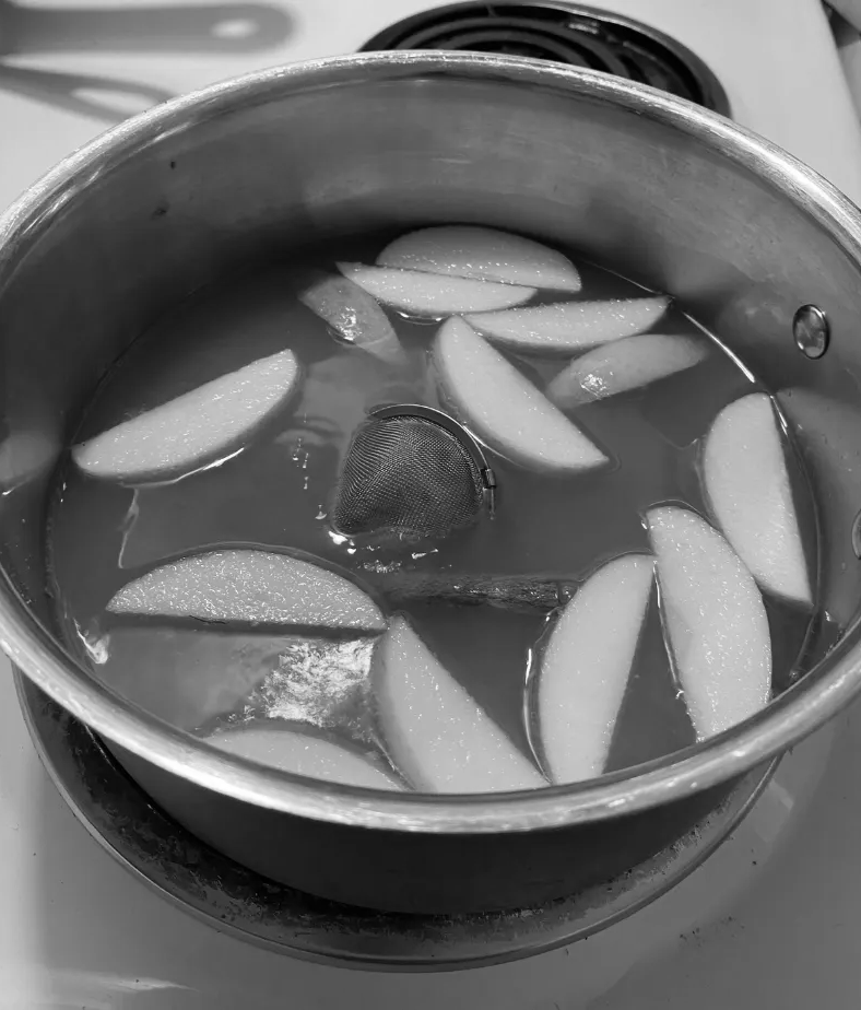 A pot on a stove filled with liquid, apple slices, and a whole spice, likely for simmering apple cider or mulled fruit.