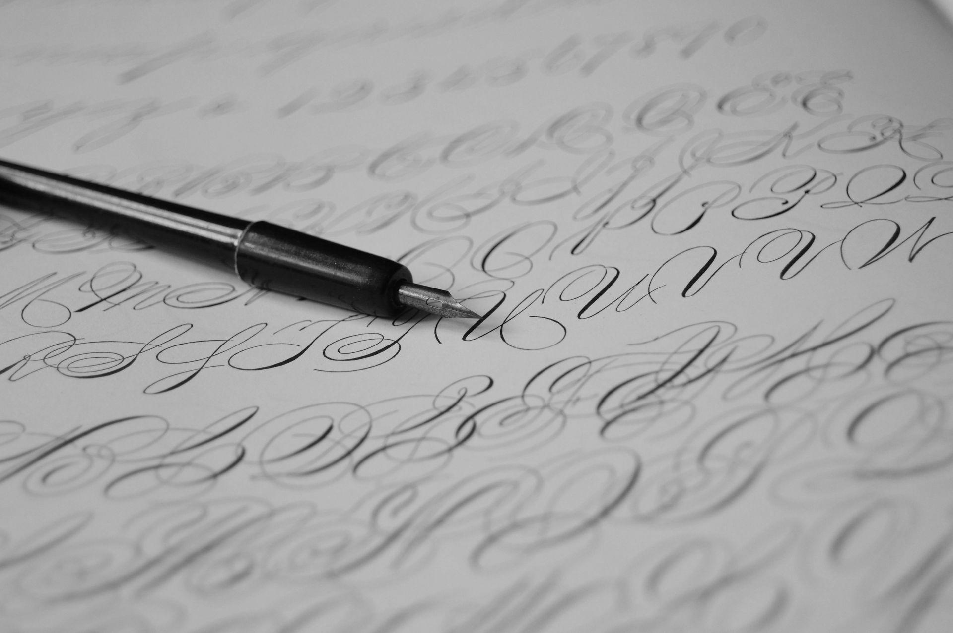 A black calligraphy pen rests on paper filled with practice loops and cursive alphabet lettering.