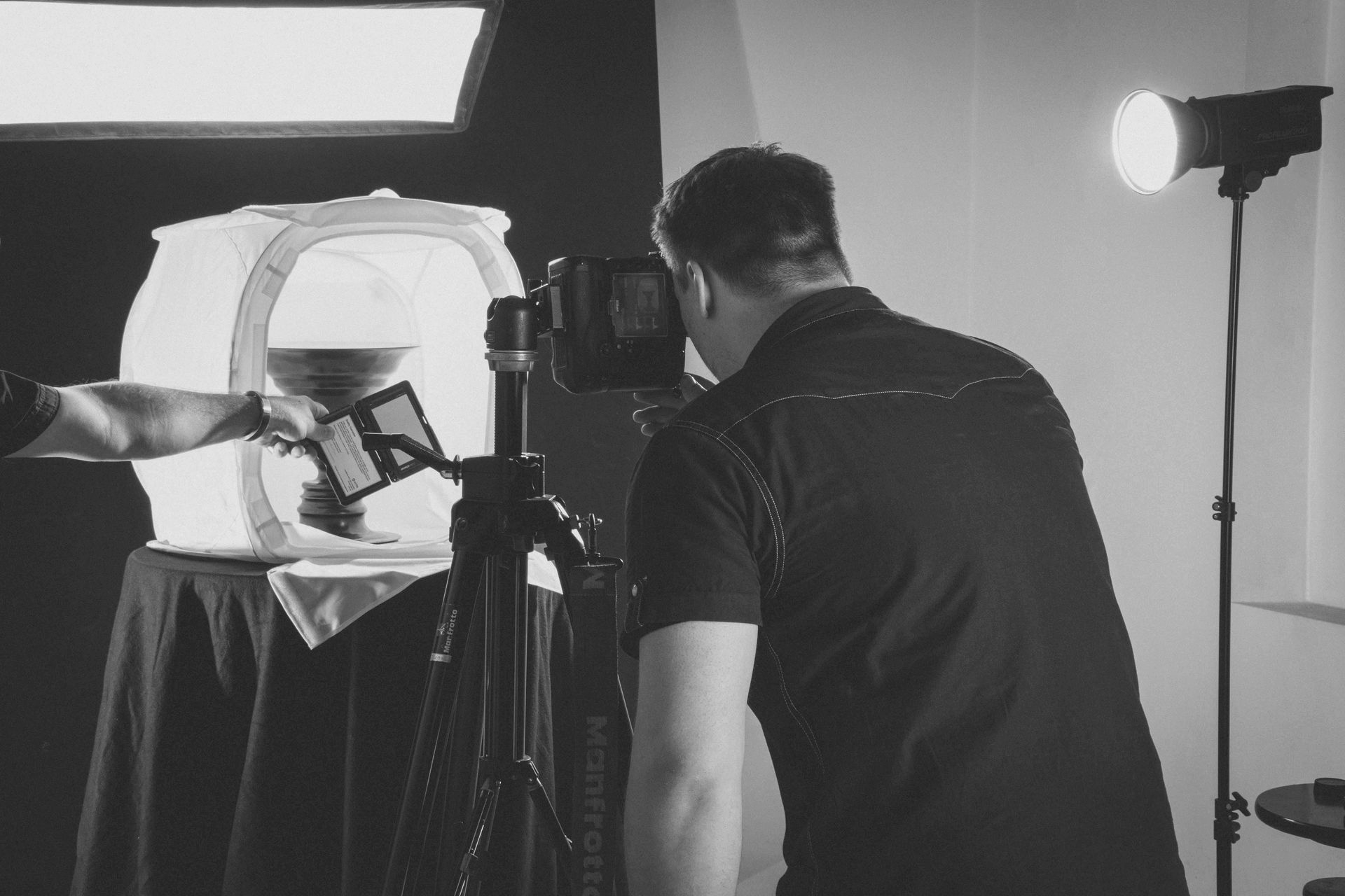 A photographer uses a camera on a tripod to photograph an object inside a light tent in a studio.