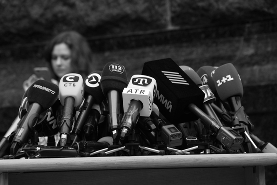 A cluster of news microphones, including 1+1 and ATR, sits on a press podium with a blurred figure in the background.