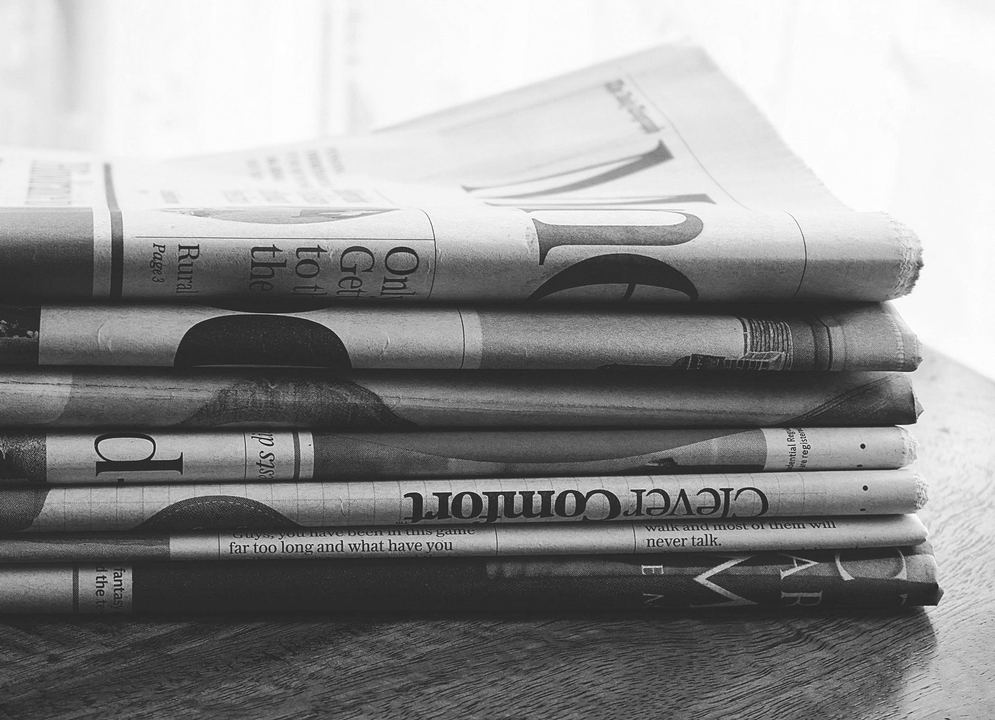 A stack of newspapers rests on a wooden surface, shown in black and white.
