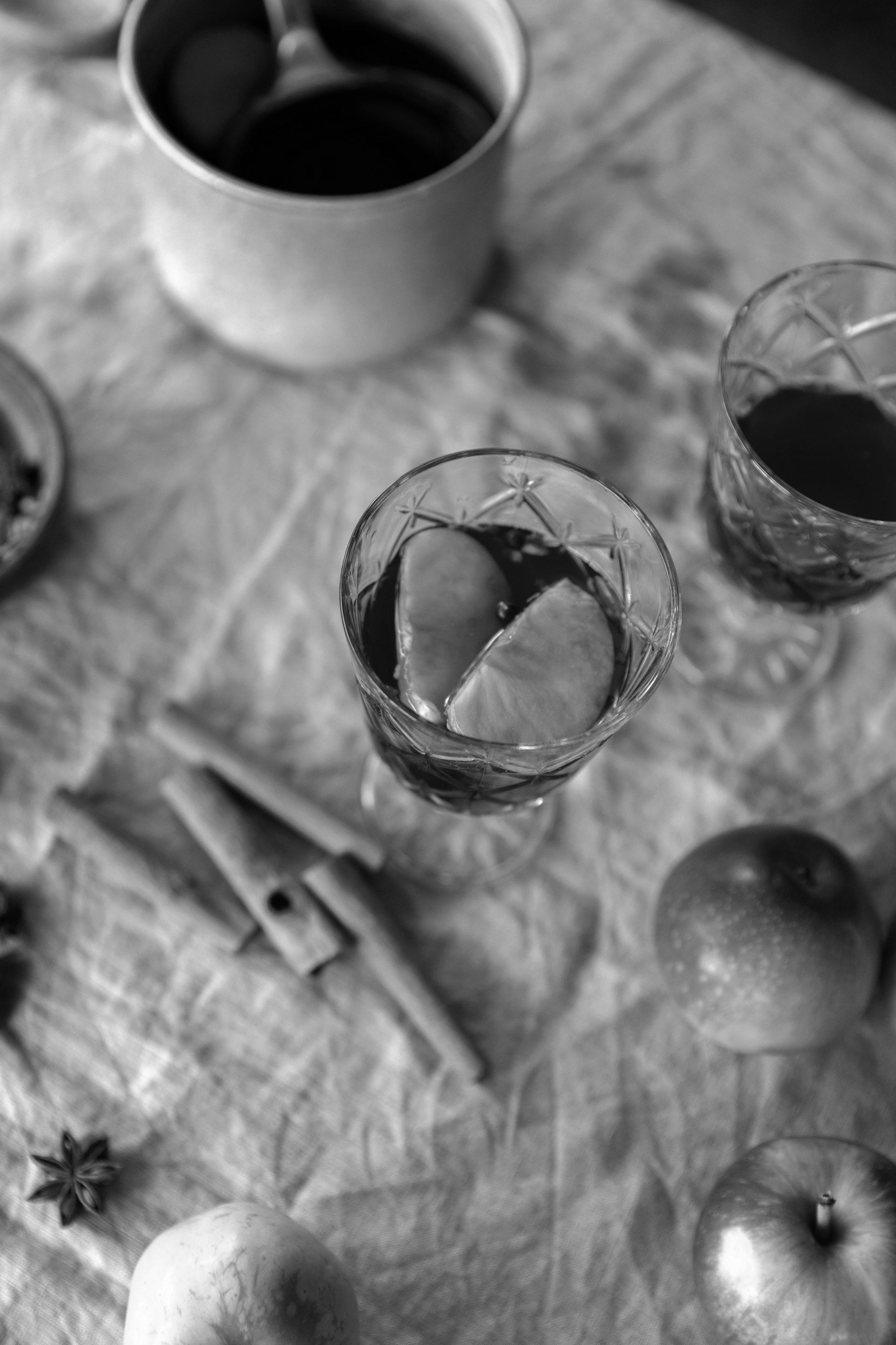A high-angle, black-and-white shot of two glasses of mulled wine with orange slices, cinnamon sticks, and apples on a cloth.