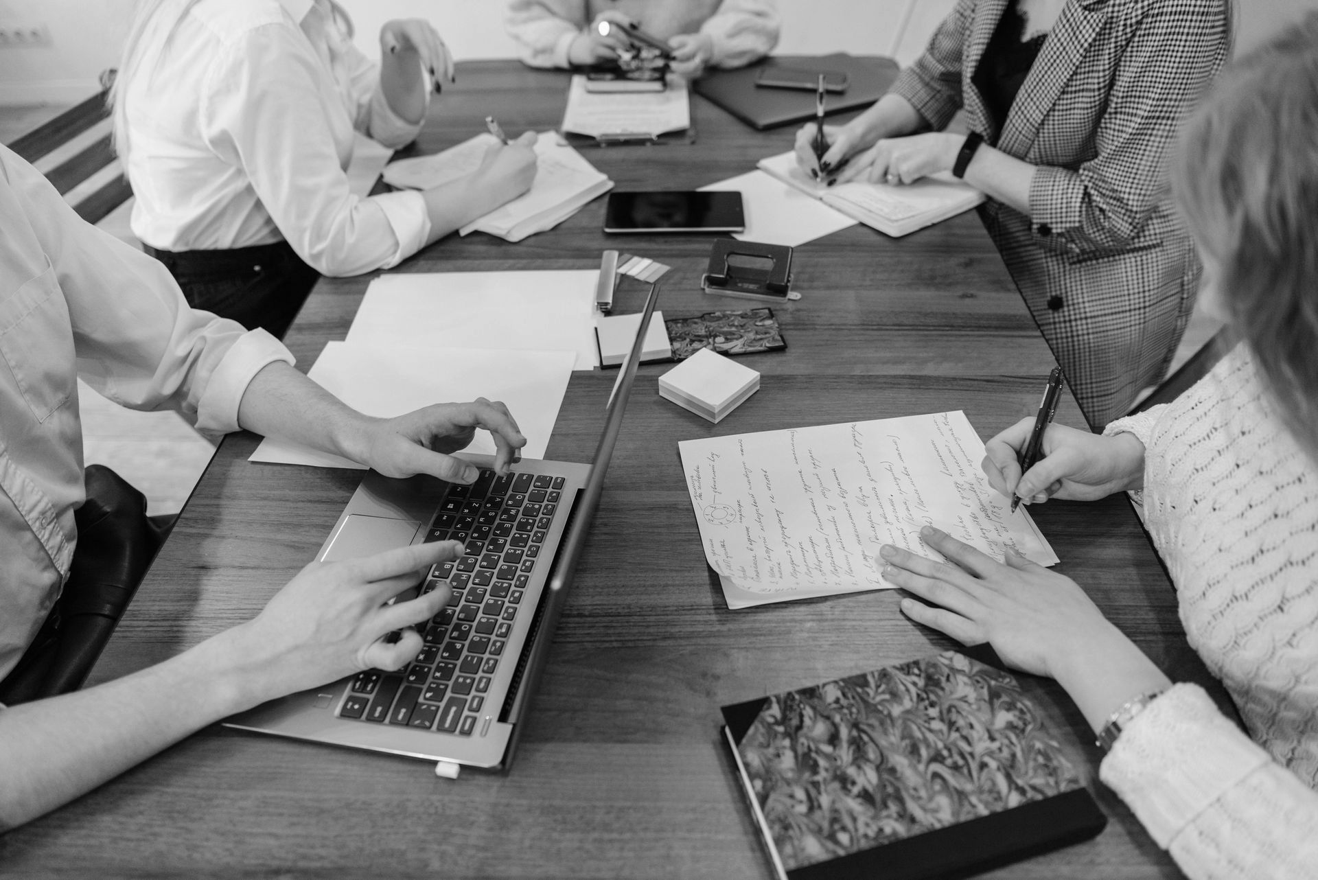 A group of people sitting around a wooden conference table working together, using laptops and writing on documents.