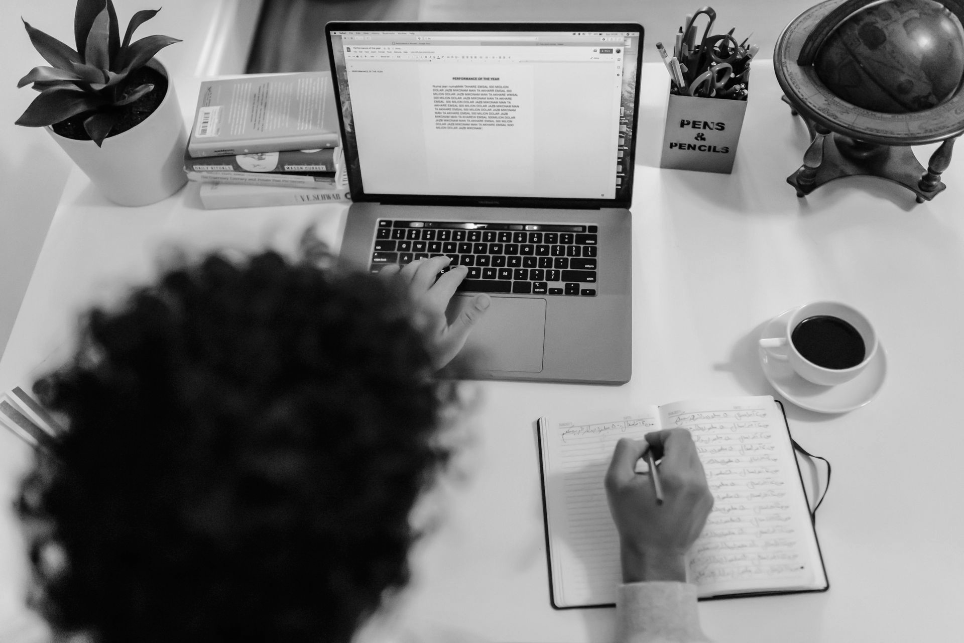 An overhead view of a desk featuring a laptop, a notebook being written in, a plant, and a coffee cup in grayscale.