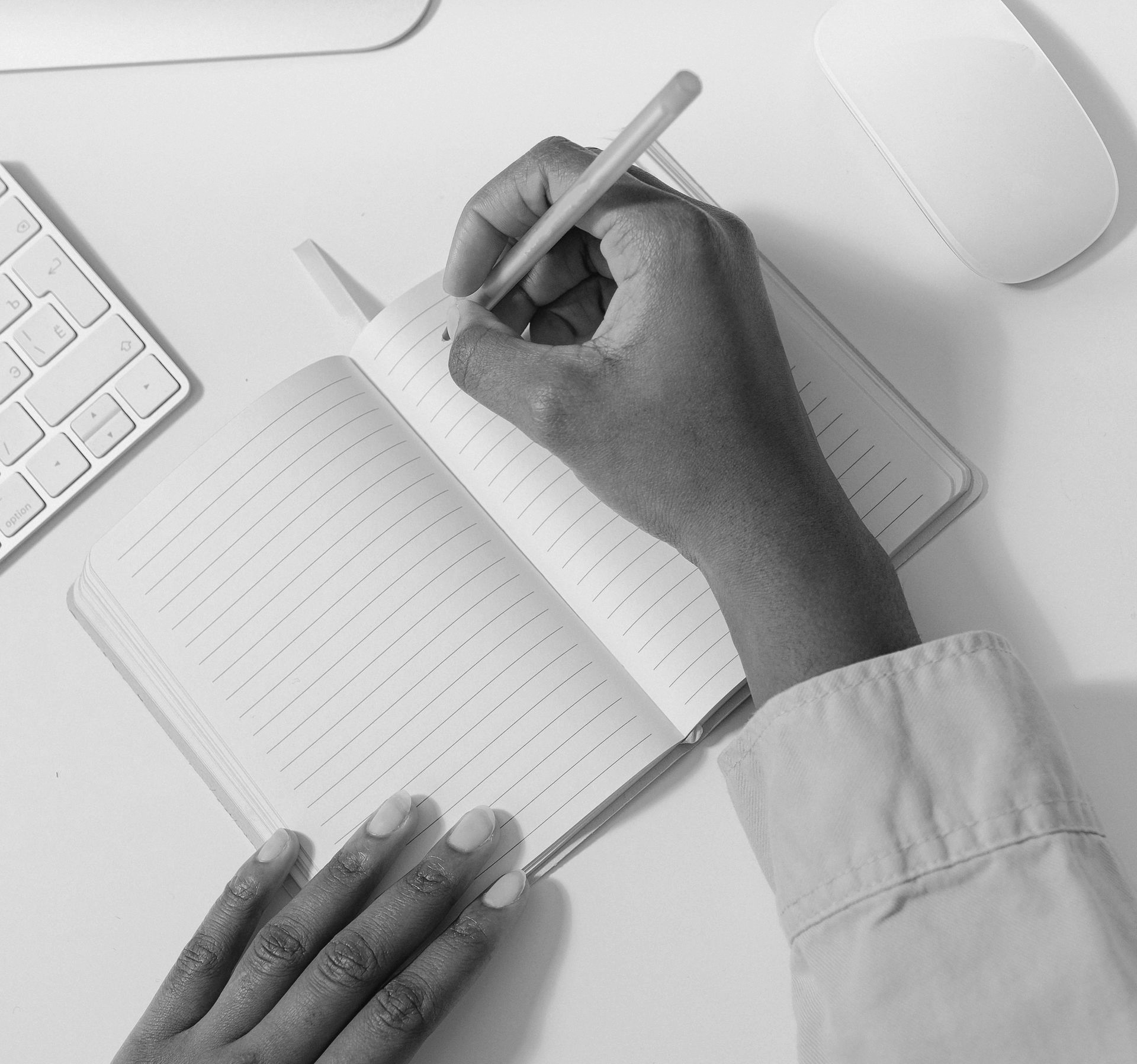 A person’s hands writing in an open notebook on a desk next to a keyboard and a computer mouse.