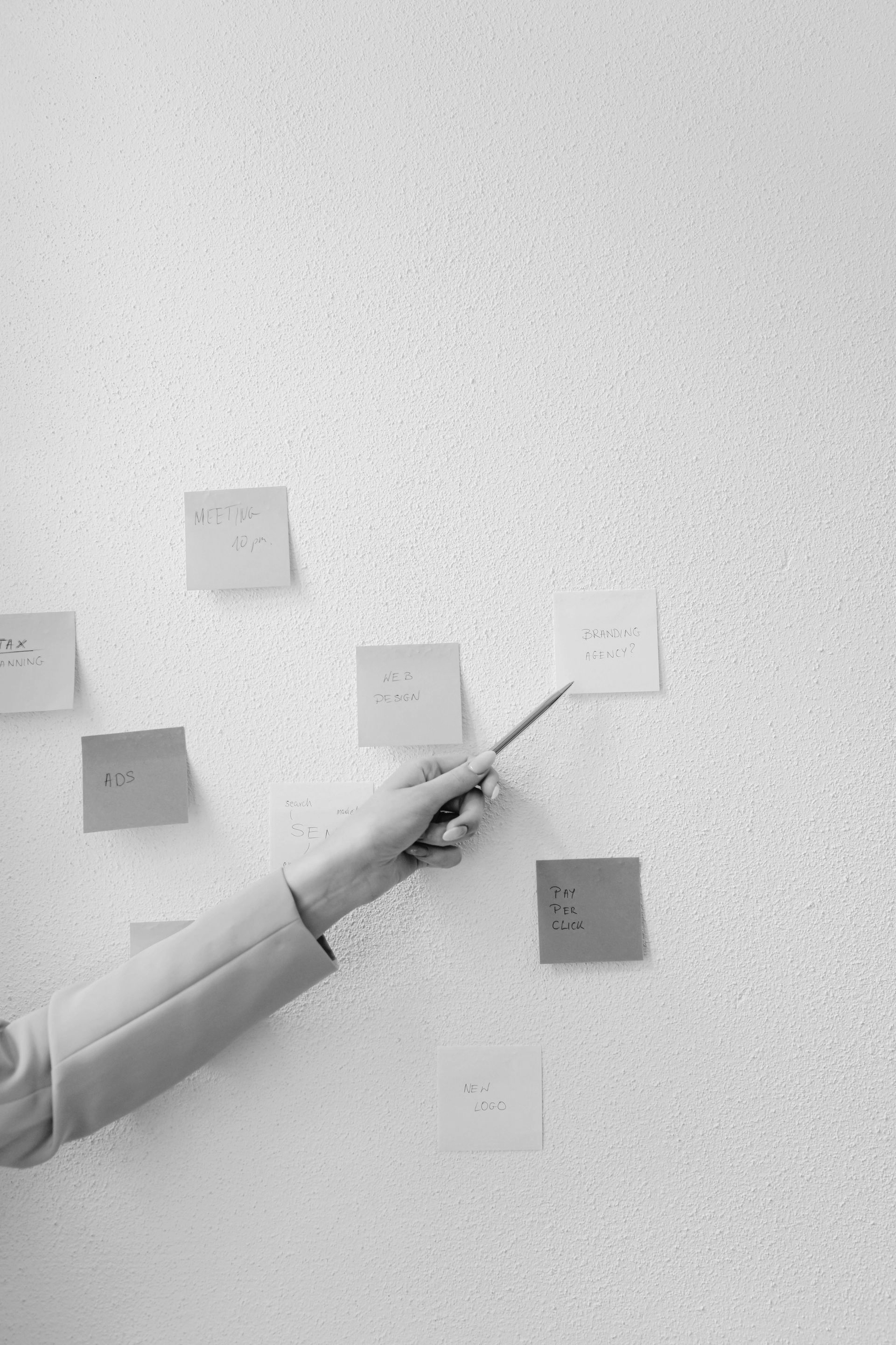 A hand in a light-colored sleeve points with a pen at a blank square sticky note on a white wall covered in sticky notes.