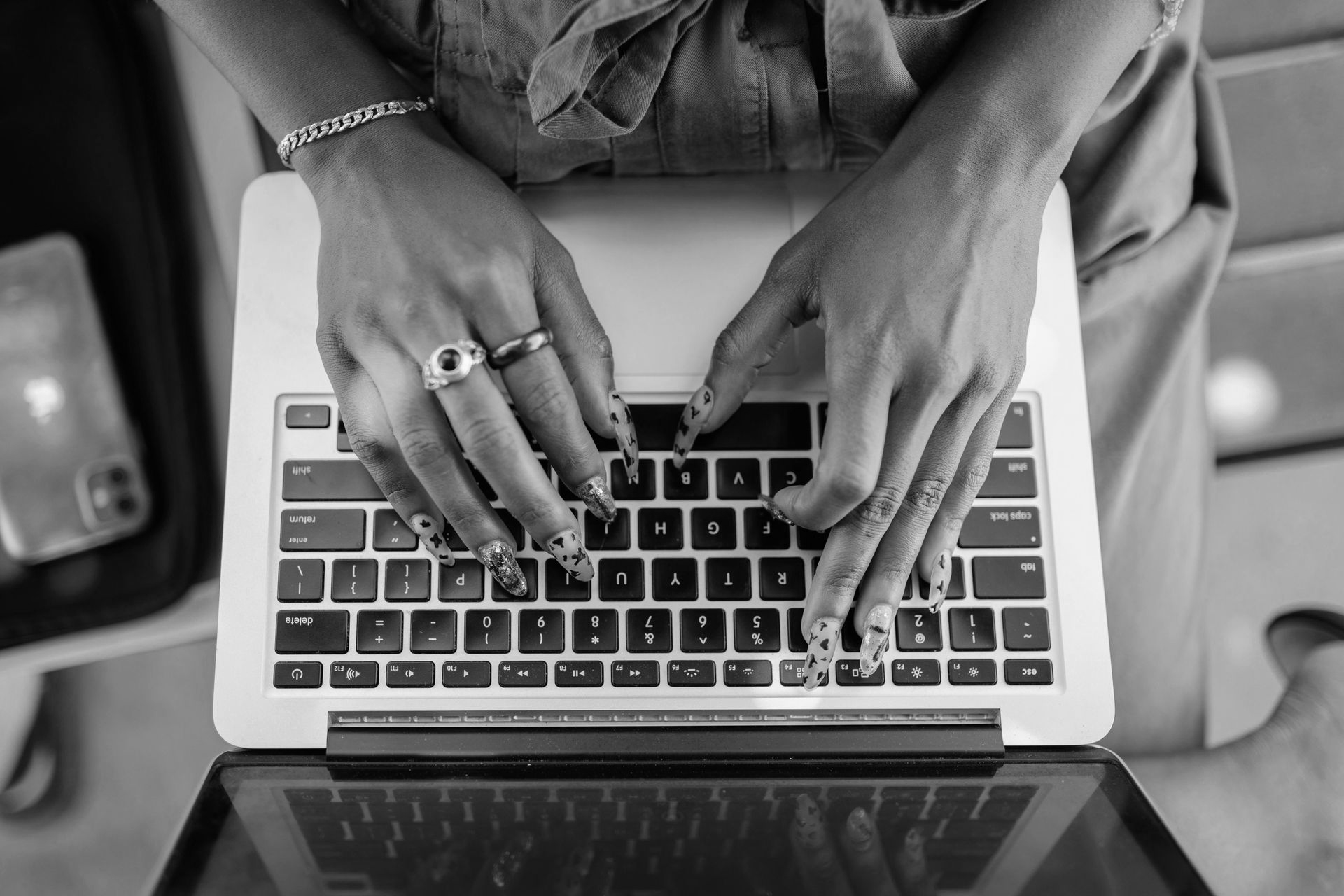 A close-up, top-down, black and white view of hands with rings typing on a laptop keyboard.