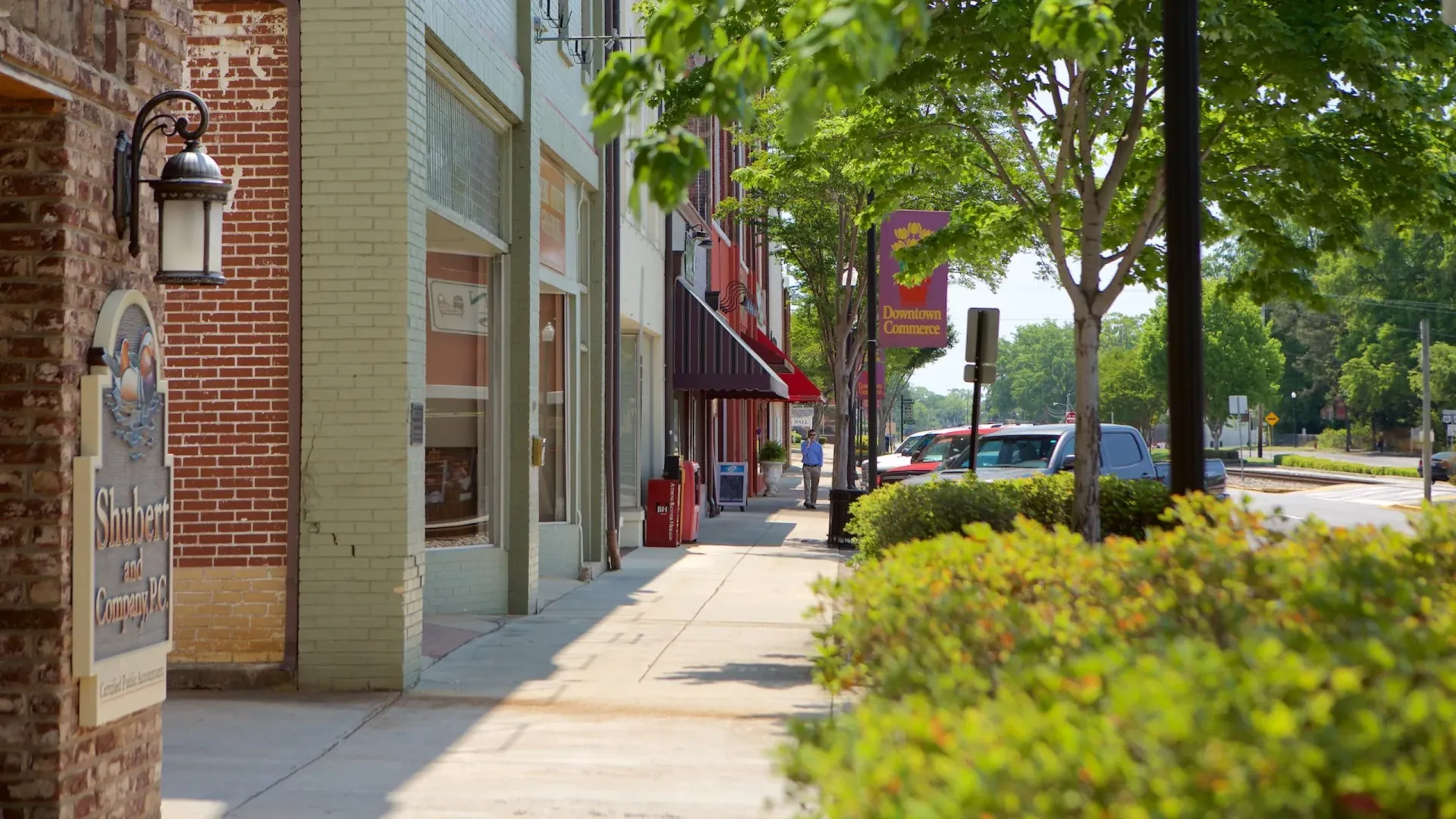 Sidewalk lined with brick buildings, shops, and trees. Sunny day.