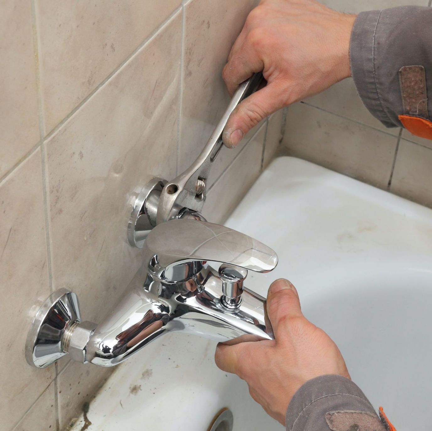 A person using a wrench to repair a silver faucet on a tiled wall next to a white bathtub. Tub leak repair in bathroom.