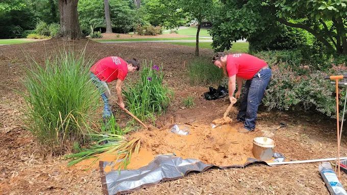MasterCraft Plumbers in red shirts are digging dirt with shovels outside, near plants and trees. Minimal yard damage plumbers