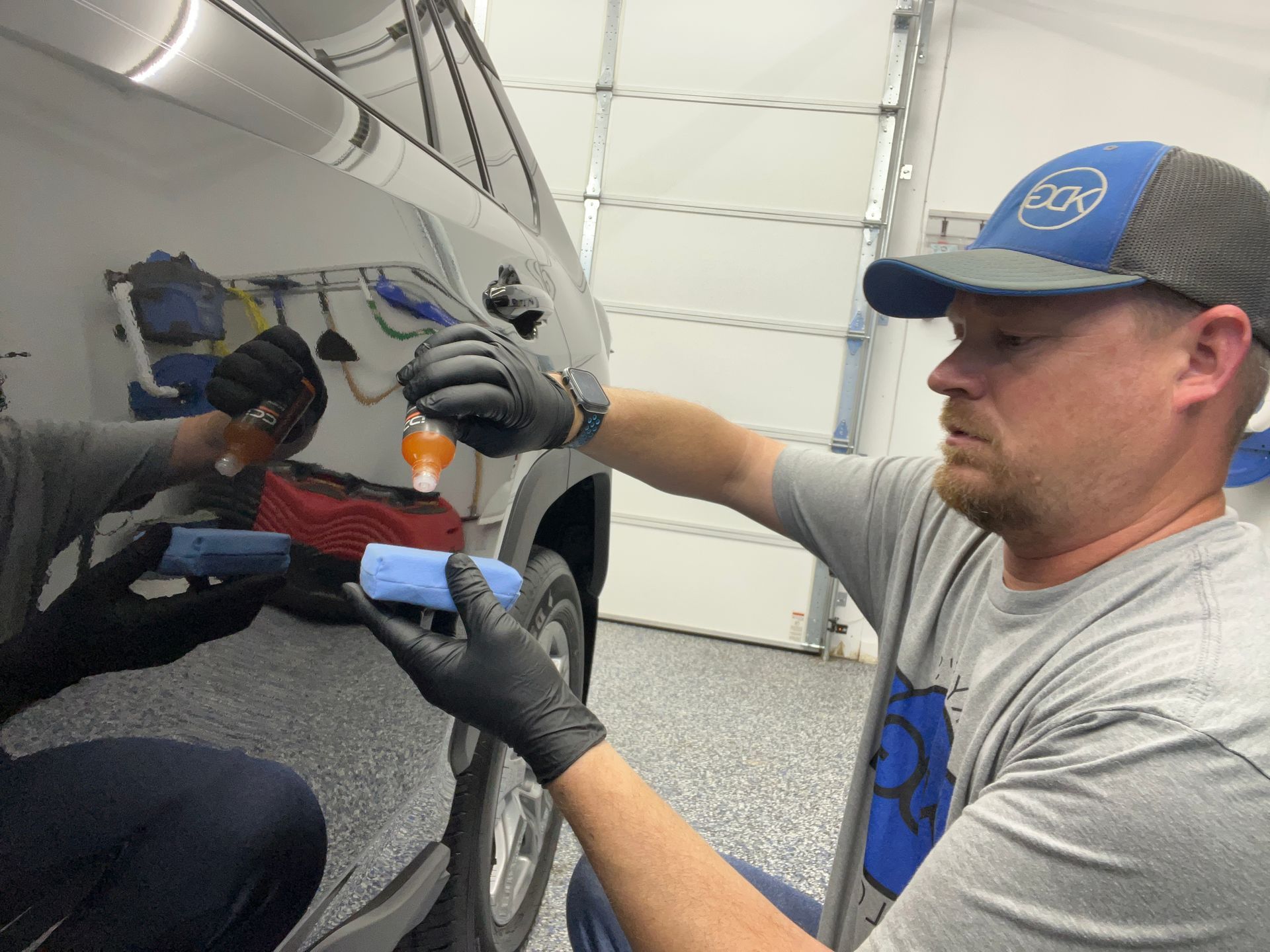 Man in cap and gloves applying a blue detailing clay bar to a vehicle's side, indoors.