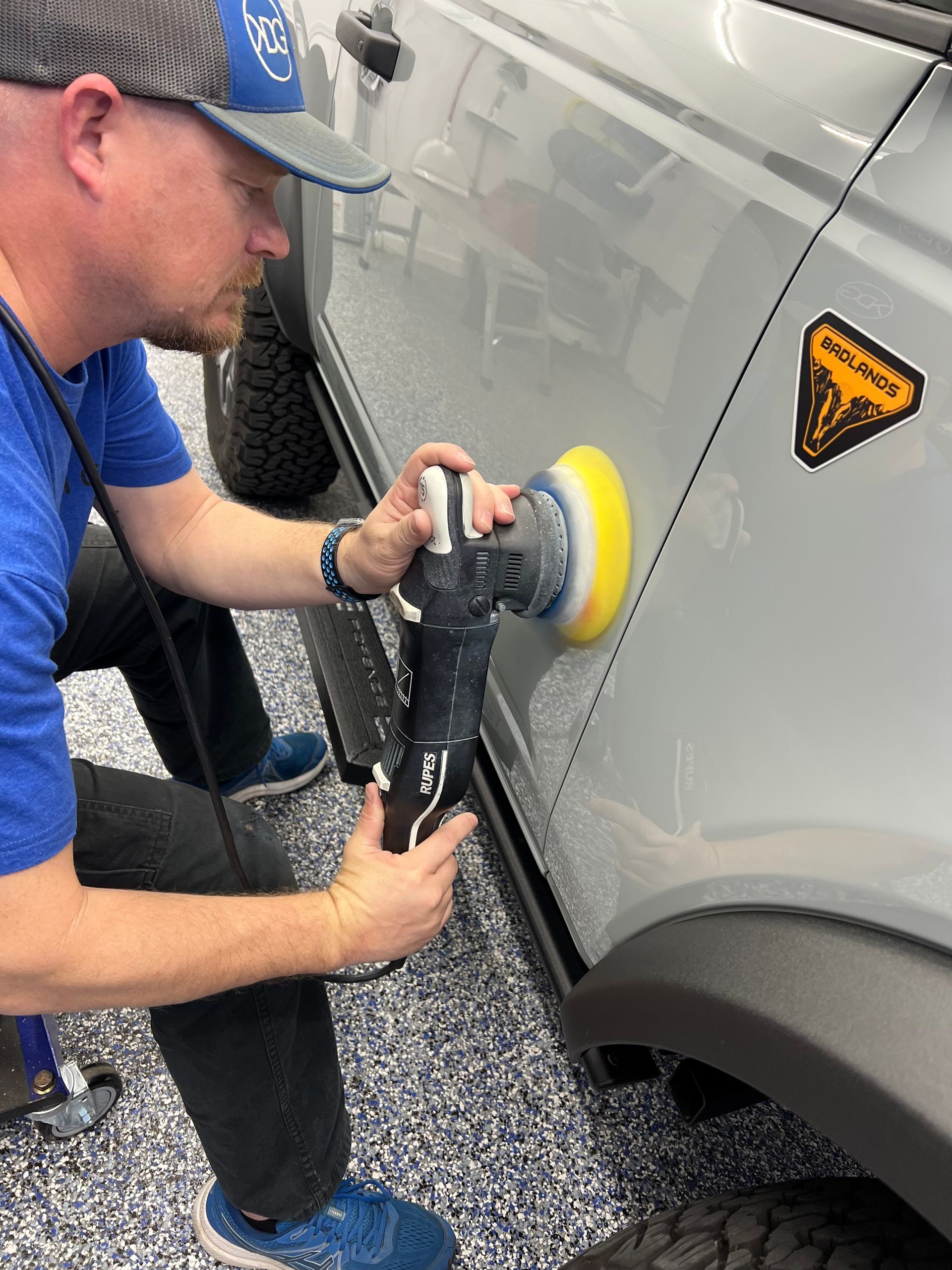 Man polishing a gray SUV with a large buffer in a garage, focusing on the vehicle's side.