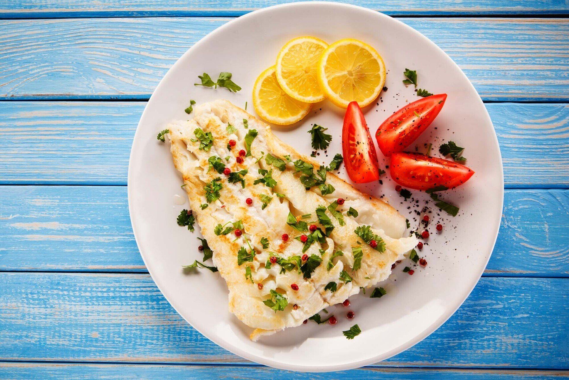 A white plate topped with fish , lemon slices and tomatoes on a blue wooden table.