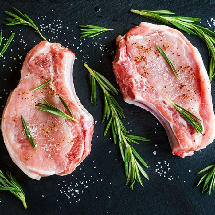 Two raw pork chops with rosemary and salt on a black surface.