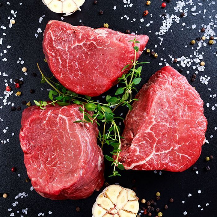Three pieces of raw beef surrounded by garlic and spices on a black surface.