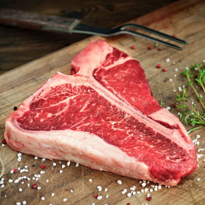 A t-bone steak is sitting on a wooden cutting board next to a fork.
