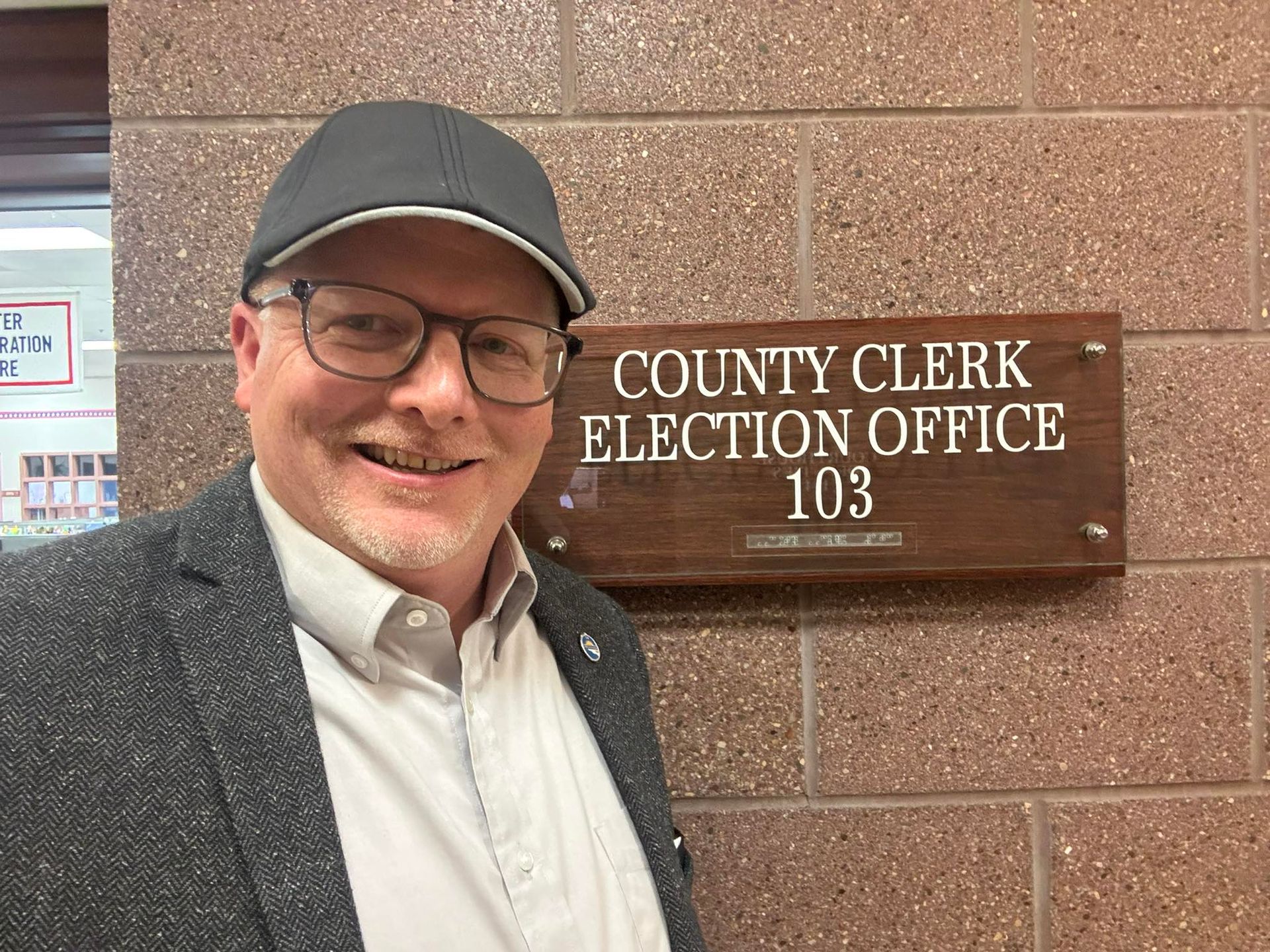 A man in a suit and hat is standing in front of a county clerk election office.