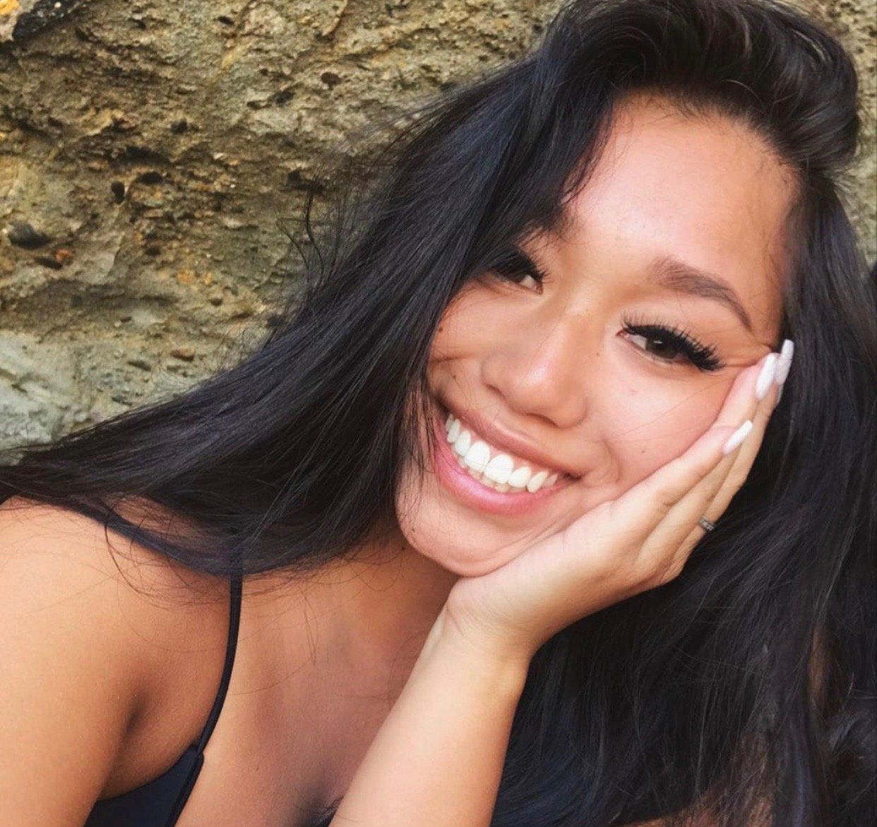 A woman with long black hair is smiling with her hand on her face