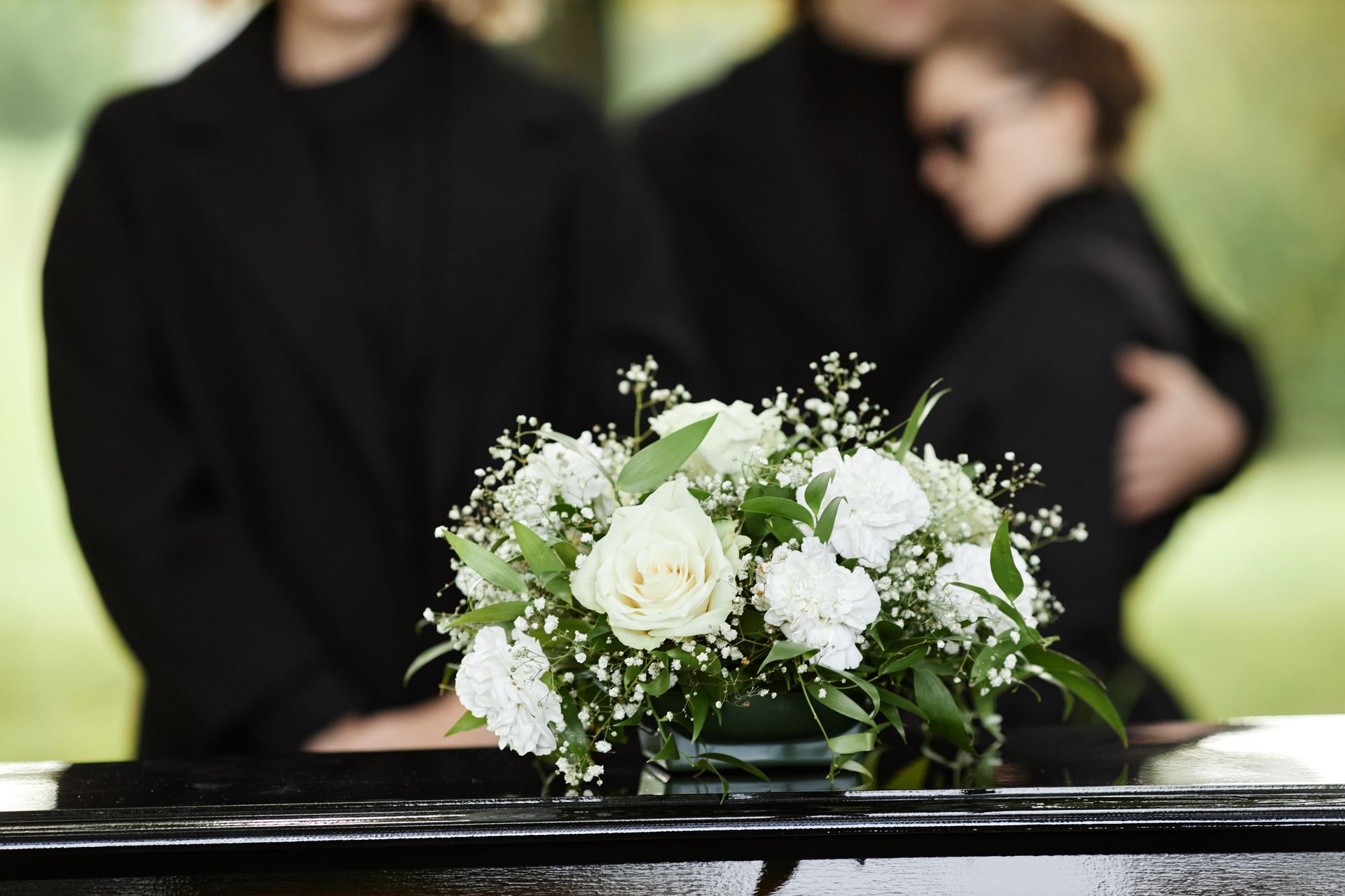 White flower arrangement on a casket at a funeral; mourners in black clothing are in the background.