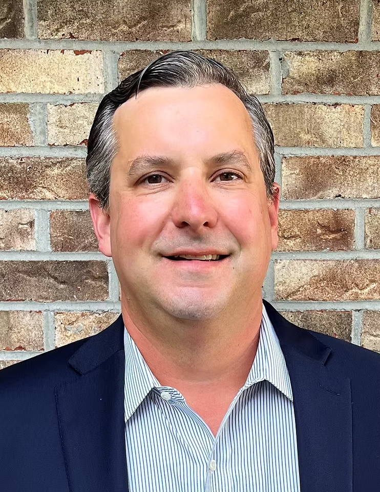 Portrait of a smiling man in a navy blazer and striped shirt against a brick wall