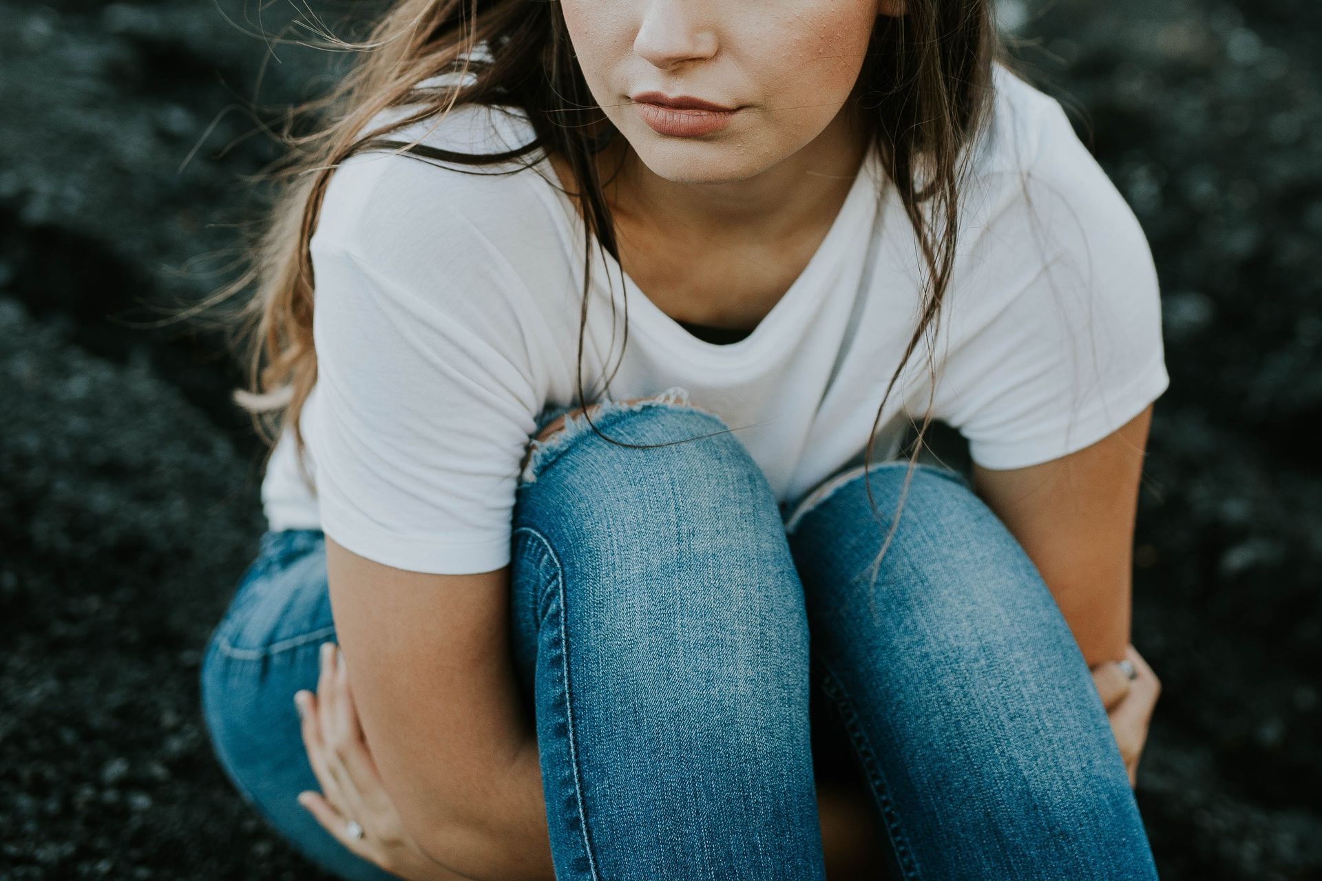 Person crouching outdoors in a white T-shirt and blue jeans on dark rocks