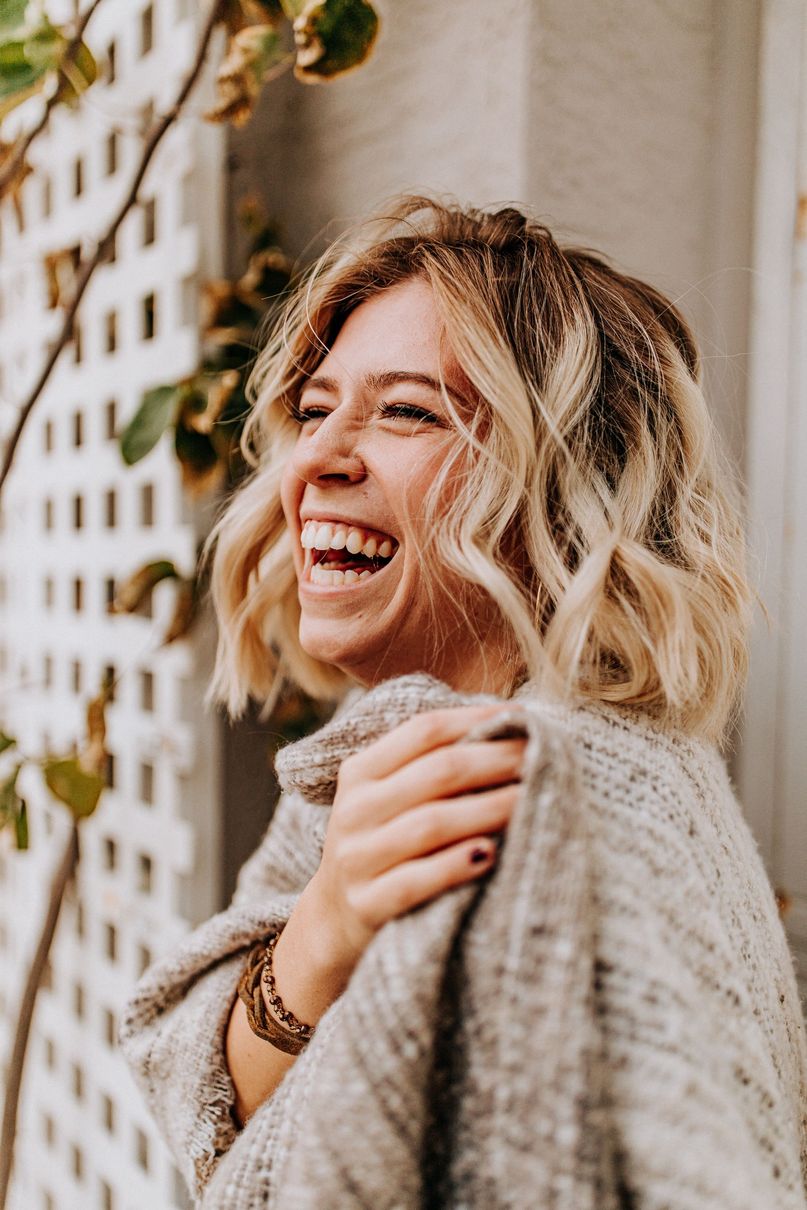 Smiling woman with wavy blonde hair in a cozy sweater, standing outdoors by a lattice wall