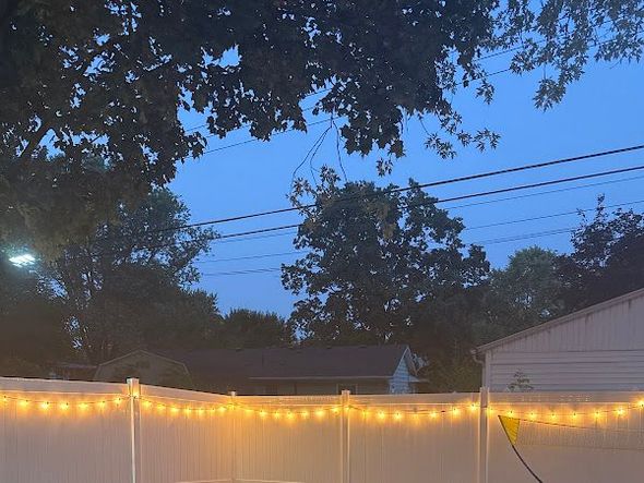 A string of warm, glowing patio lights draped along a white vinyl fence against a twilight sky with trees in the background in  Columbus