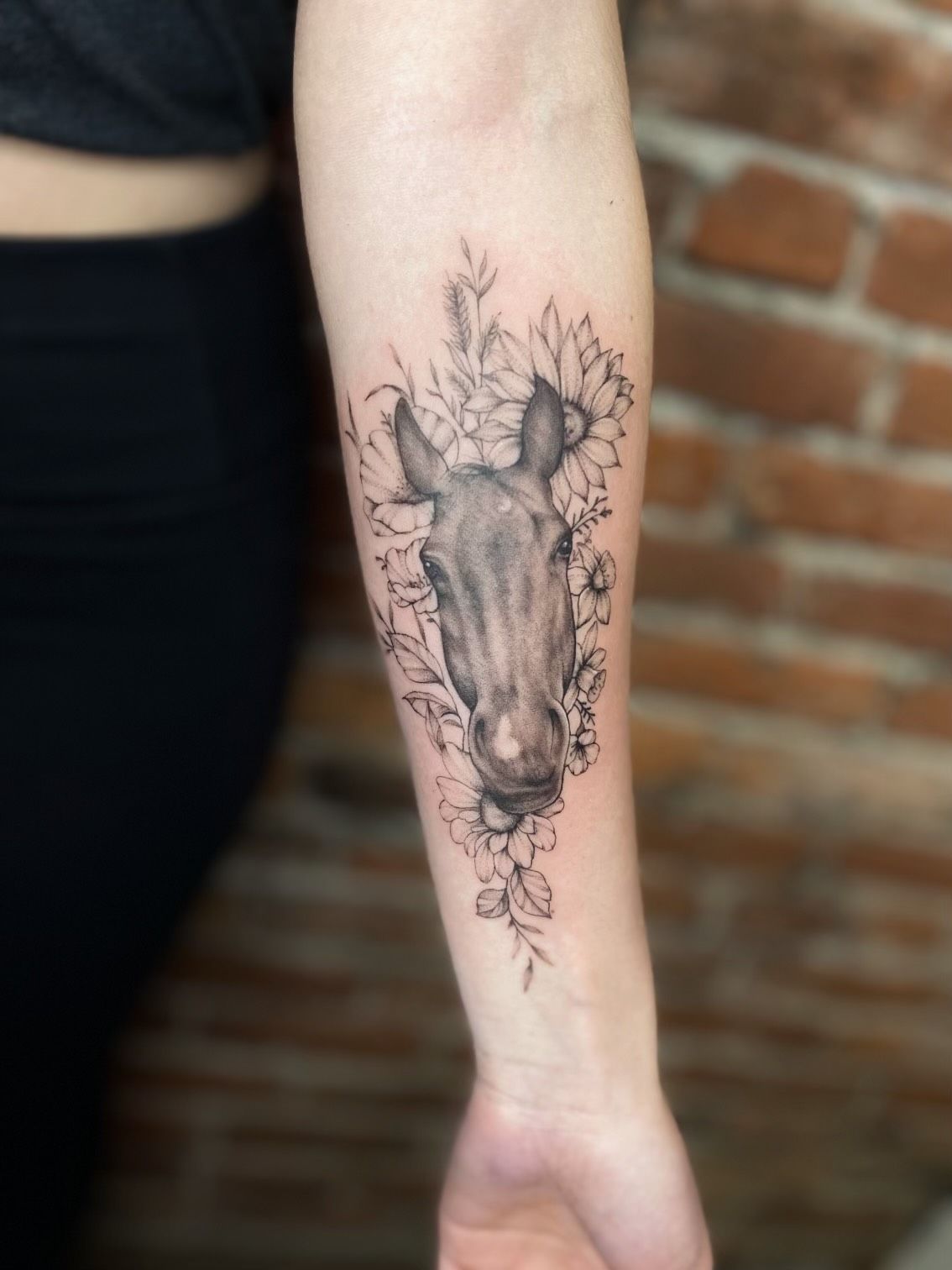 A woman has a tattoo of a horse 's head surrounded by flowers on her forearm.