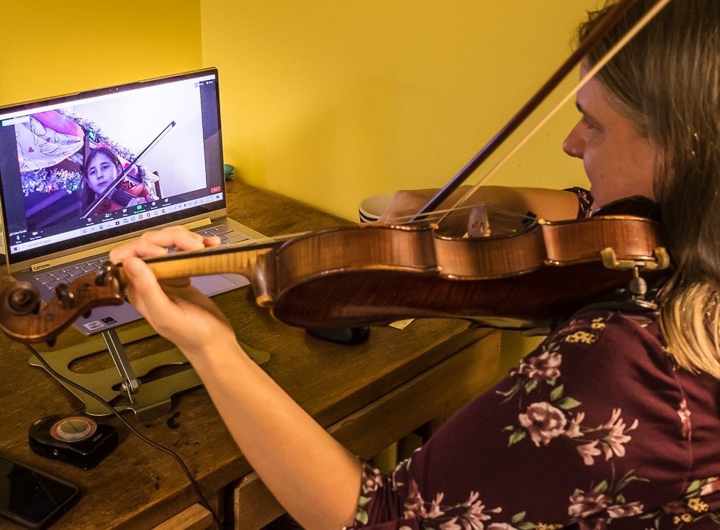 A violin teacher wearing a floral top gives an online lesson to a student visible on a laptop screen.