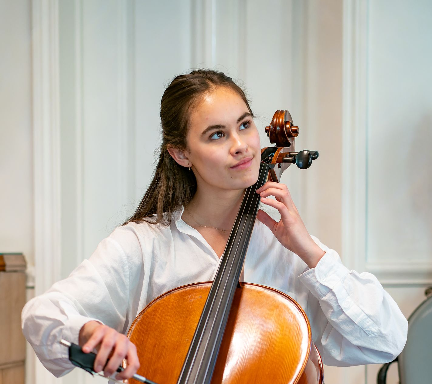 A person wearing a white shirt holds a cello and bow, looking thoughtfully to the side against a white wall background.