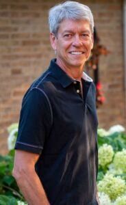 A person with grey hair smiles, wearing a dark polo shirt while standing outdoors near a brick wall and green foliage.