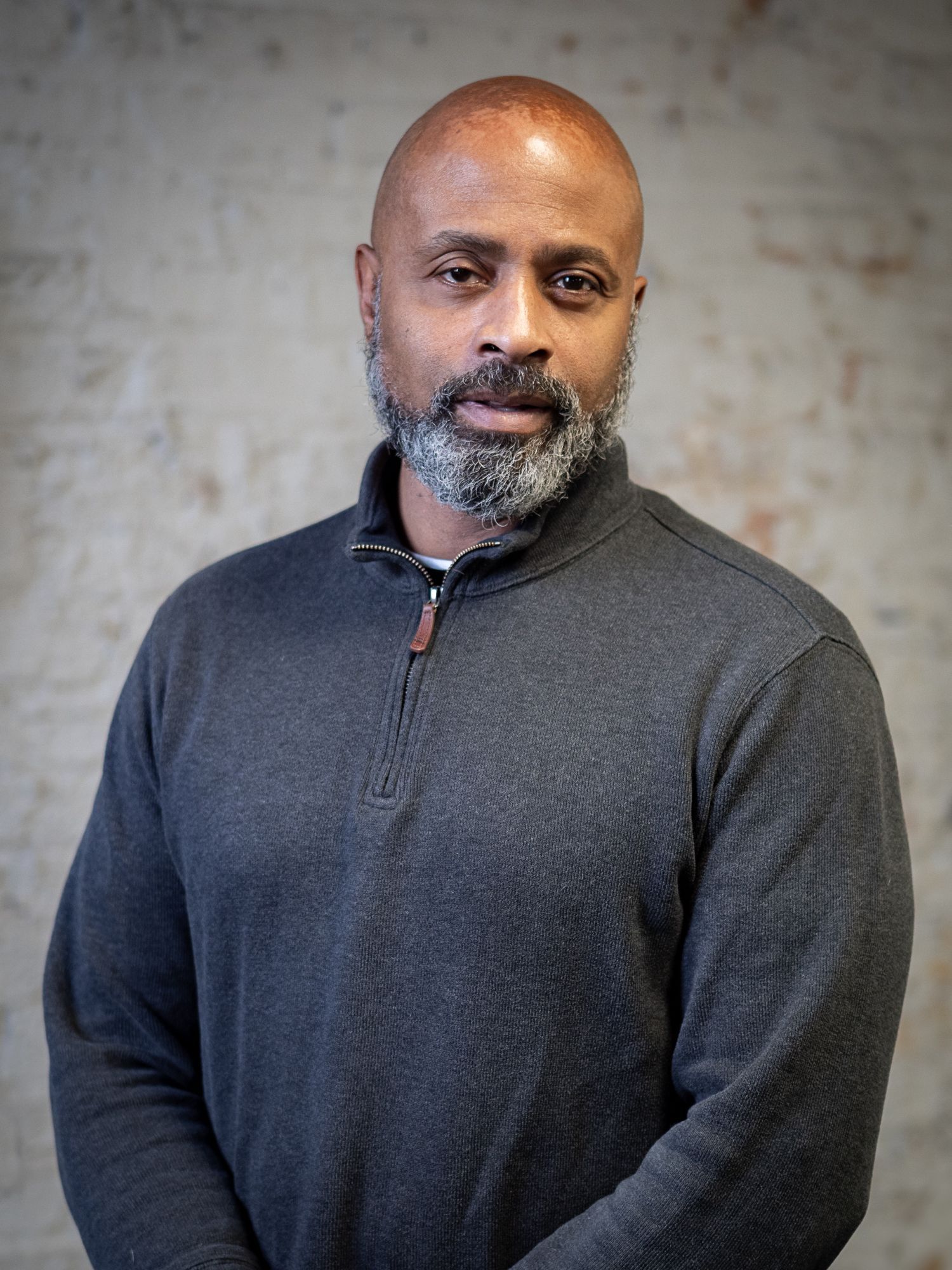 A bald man with a graying beard, wearing a charcoal gray quarter-zip sweater, stands against a textured wall.