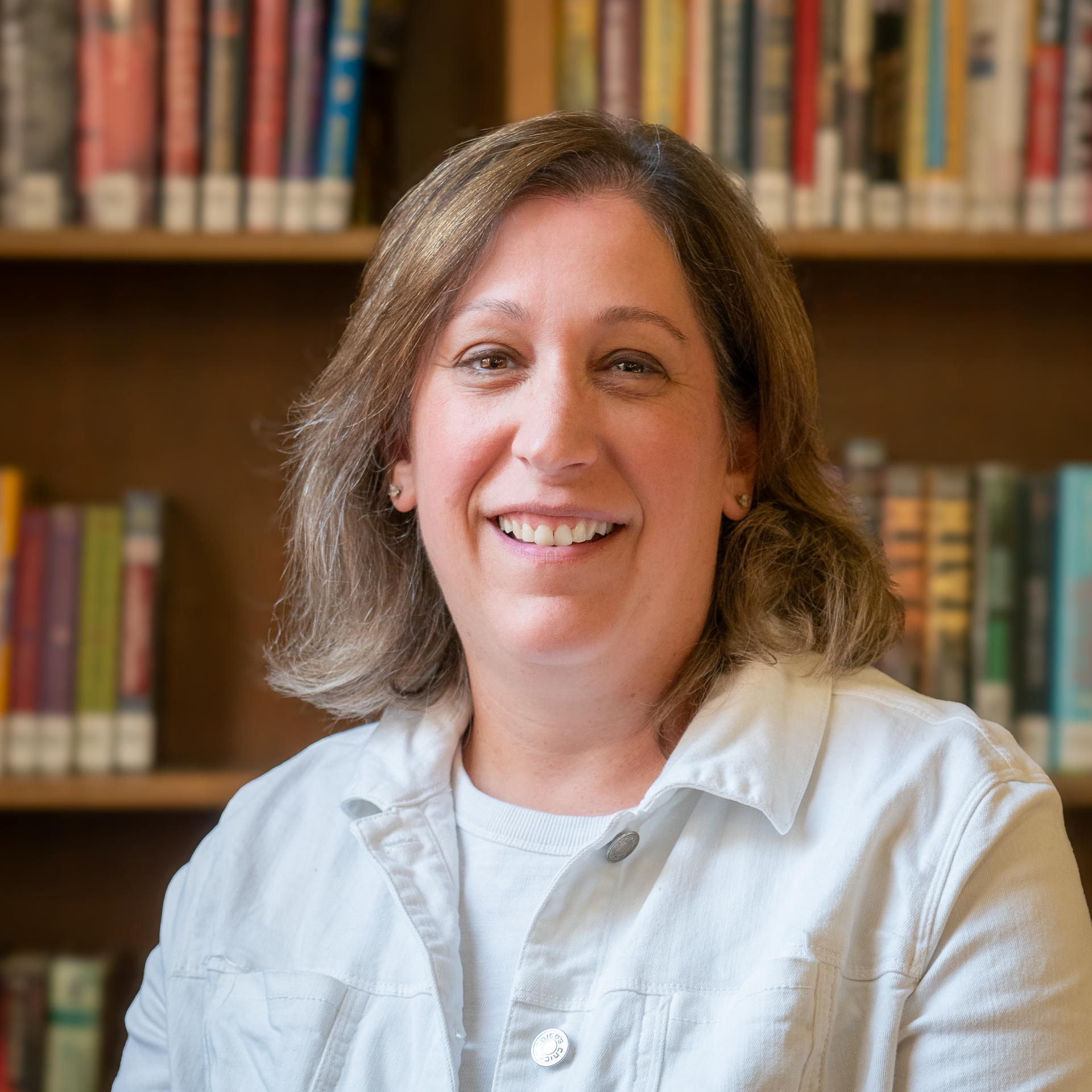A smiling person wearing a white jacket stands in front of bookshelves.