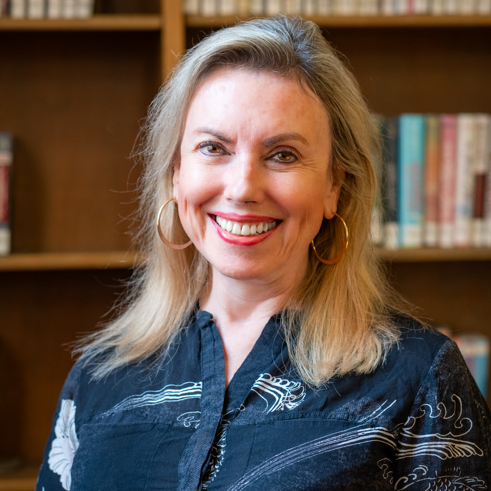 A smiling woman with blonde hair and hoop earrings, wearing a dark patterned shirt, standing in front of bookshelves.