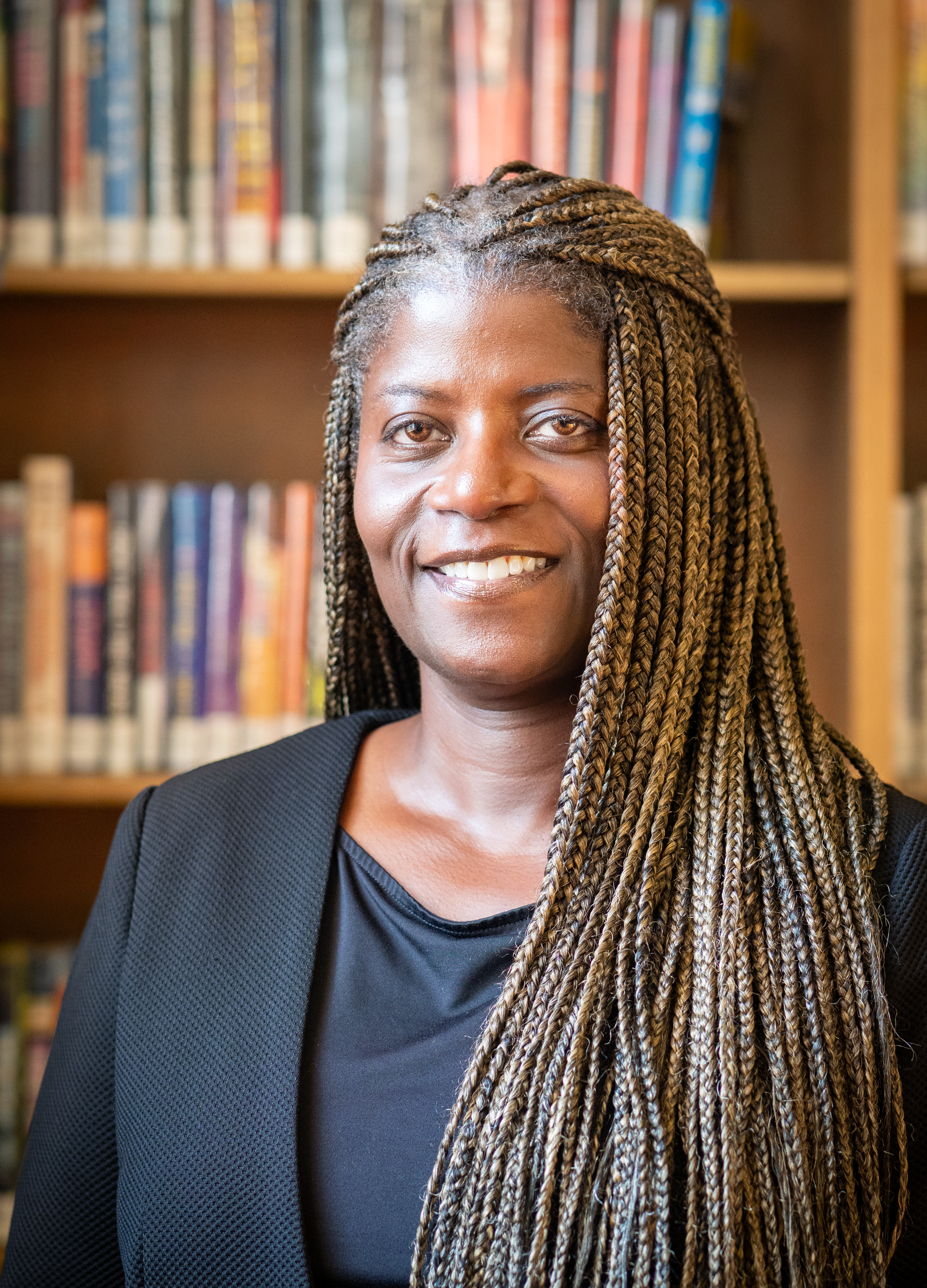 A smiling individual with long braided hair wearing a dark blazer, positioned in front of a library bookshelf.