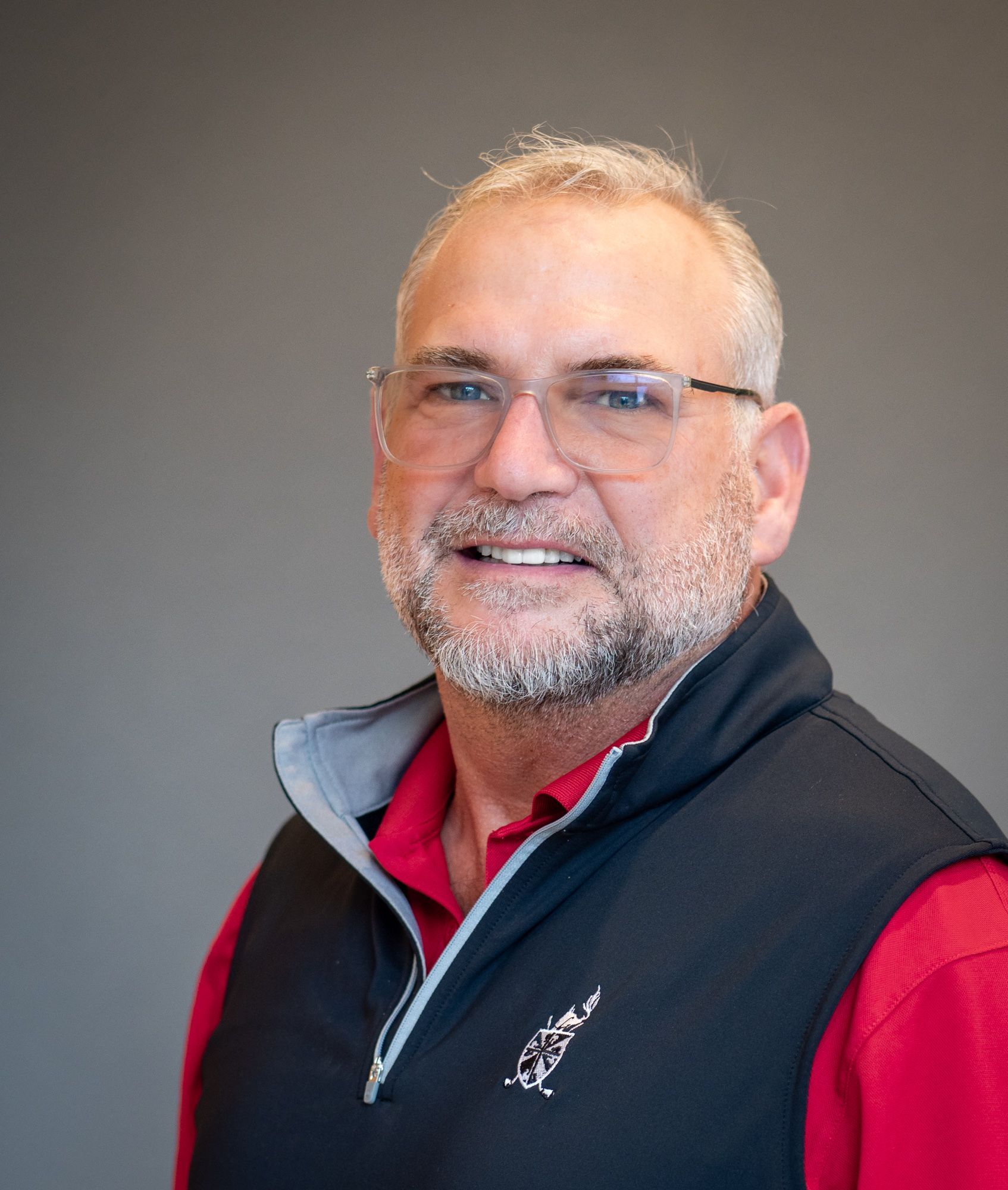 A smiling person with glasses, a gray beard, and a red shirt under a dark blue quarter-zip vest against a gray background.