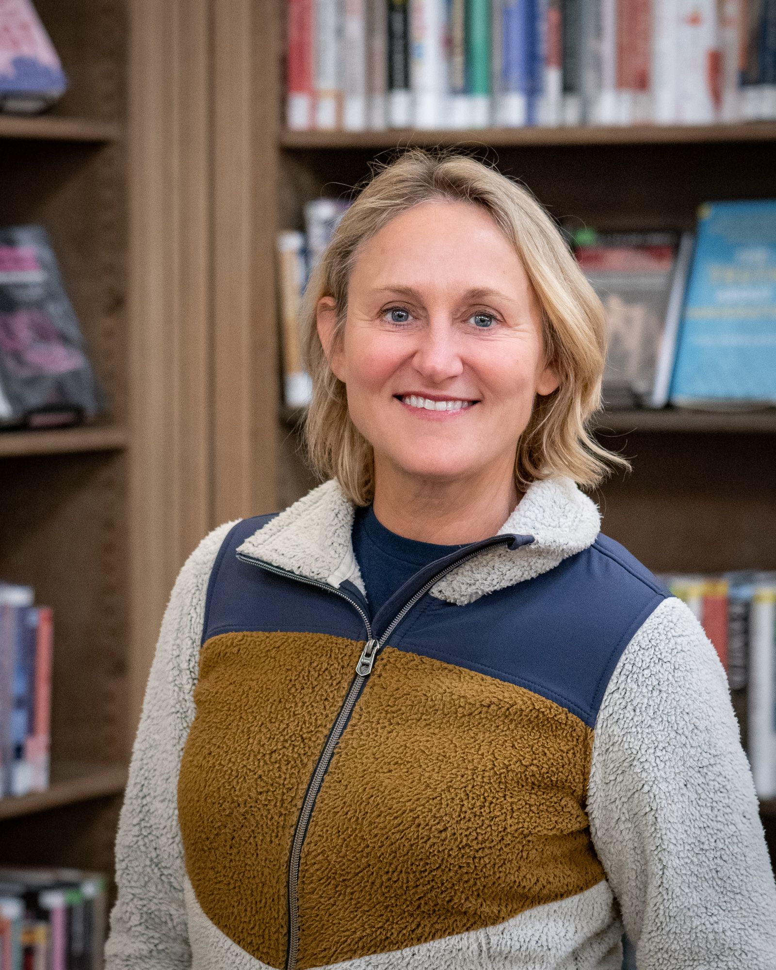 Smiling person wearing a mustard and navy fleece jacket, standing in front of a bookshelf filled with books.