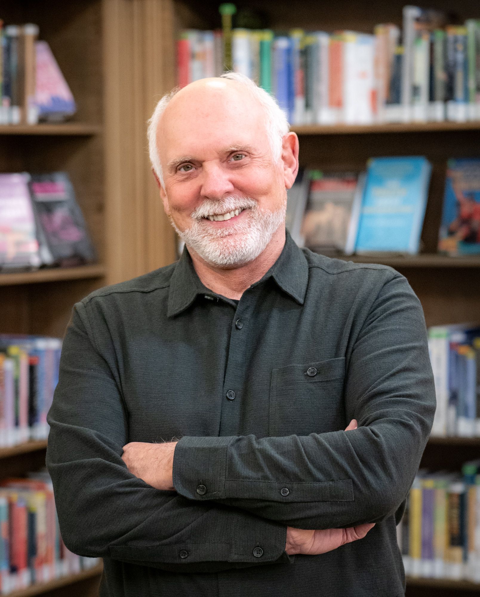 A person with a white beard and hair smiles while standing with arms crossed in front of a bookshelf.