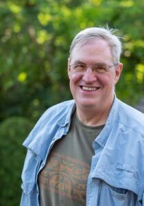 A smiling person wearing glasses, a green patterned shirt, and an unbuttoned blue overshirt, standing outdoors.