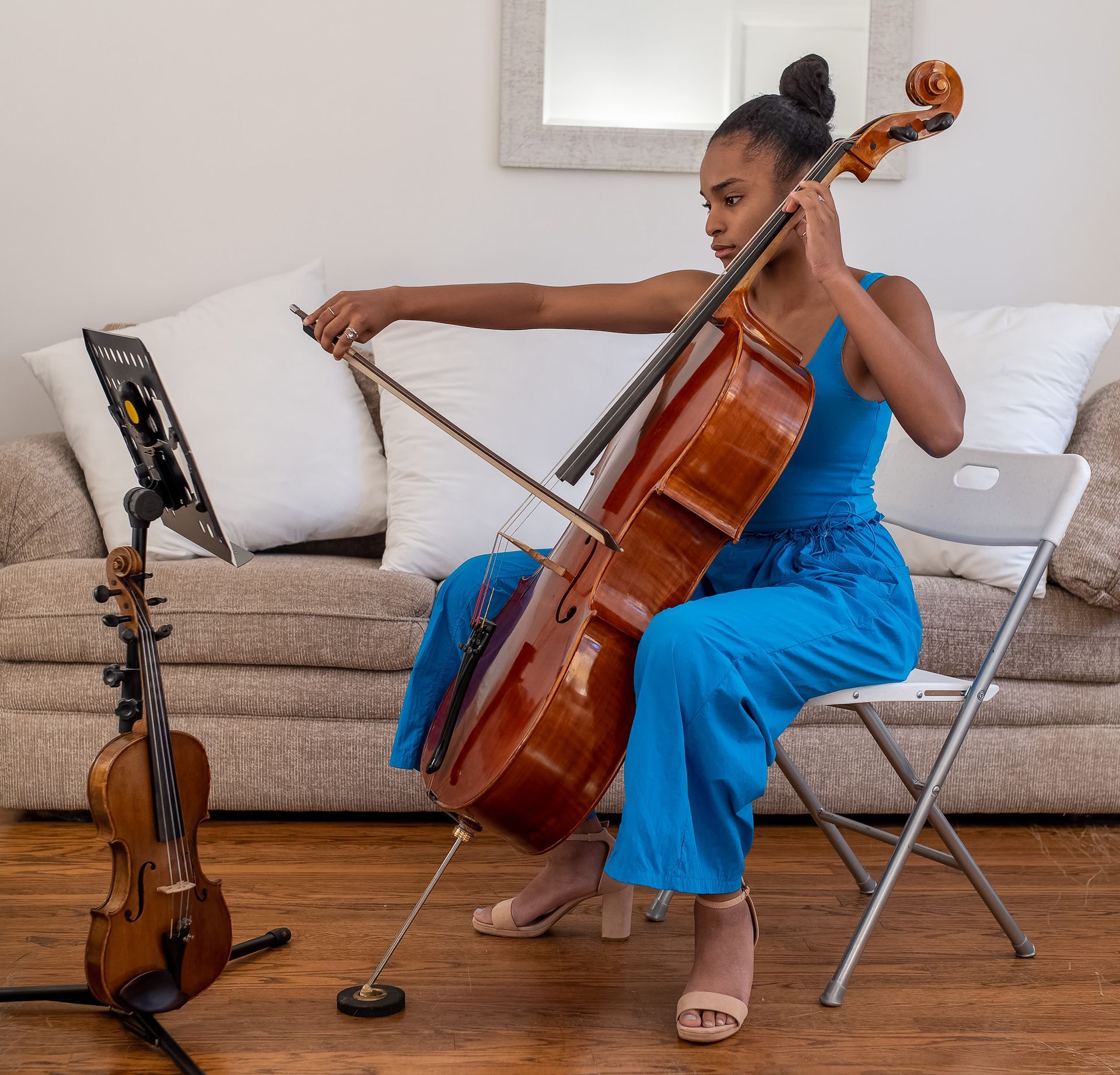 A musician in a blue outfit plays a cello while seated in front of a sofa; a violin rests on a stand to the left.