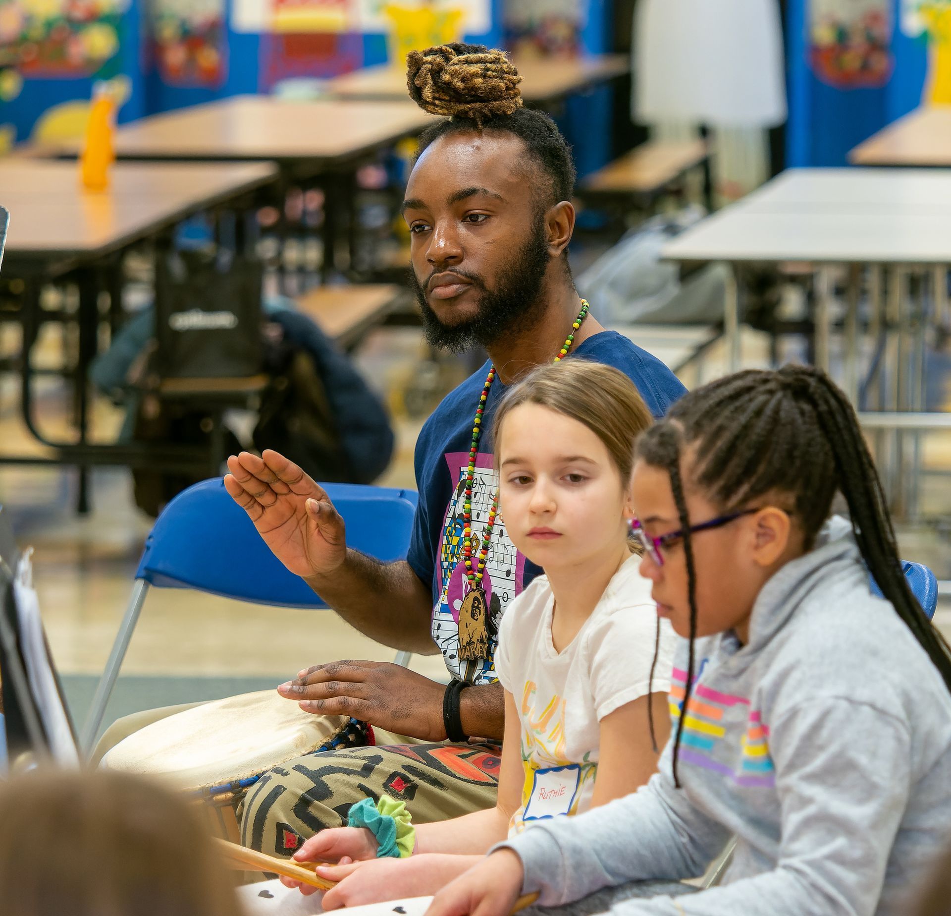 A music instructor with styled hair teaches two students to play drums in a classroom setting.