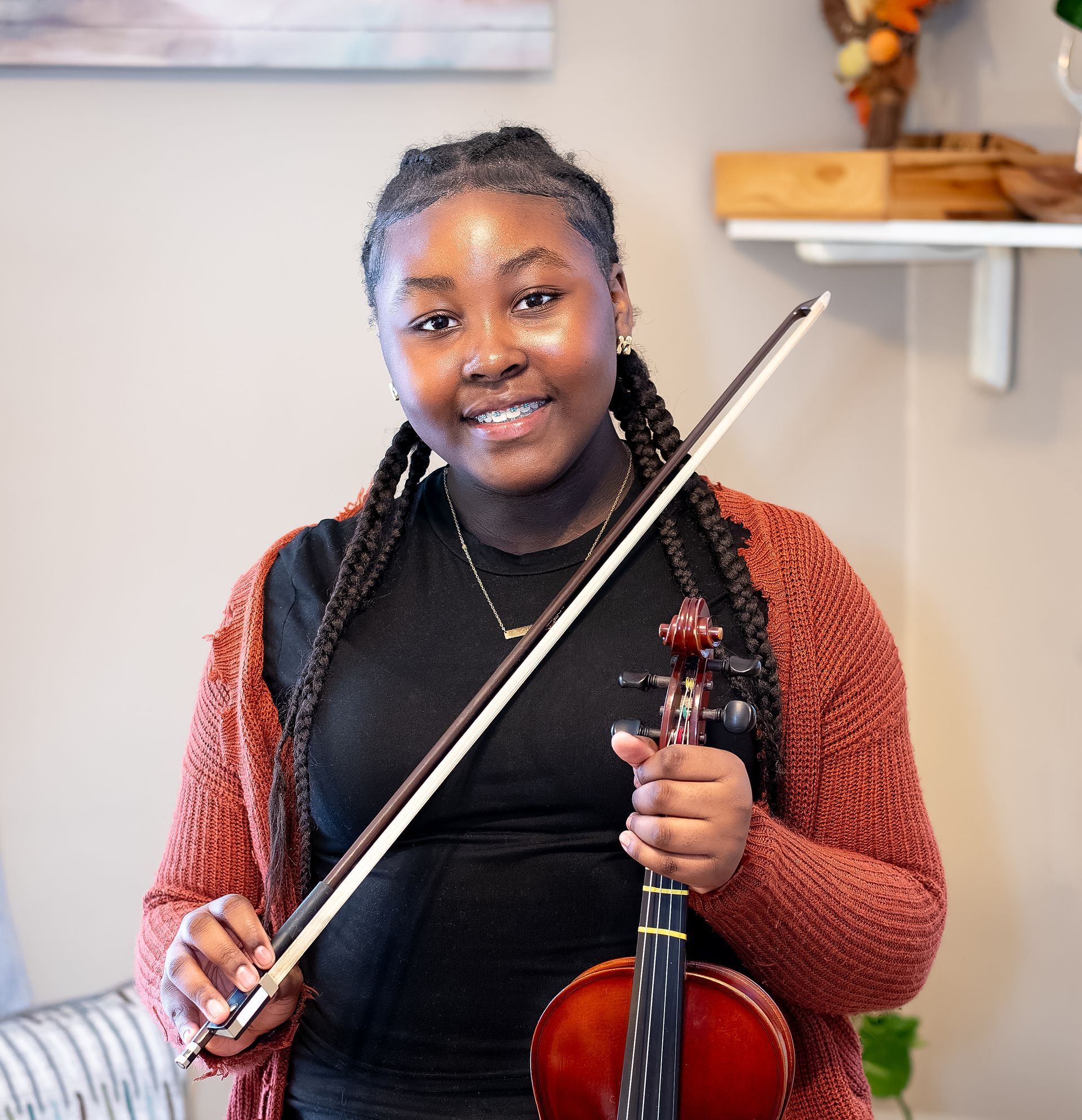 A person in an orange cardigan smiles while holding a violin and bow in an indoor room.