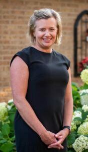 A smiling person with blonde hair in a black dress stands outdoors in front of a brick wall and green hydrangeas.