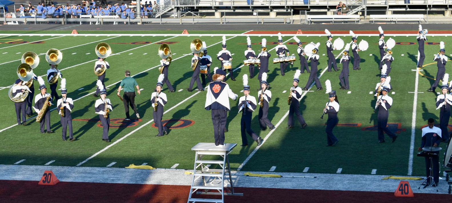 A marching band performs on a green football field, led by a conductor standing on an elevated podium.